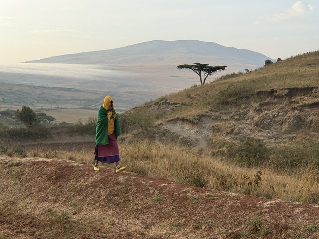 Massai in traditioneller Kleidung auf einem Pfad im Hochland, dahinter eine einzelne Akazie